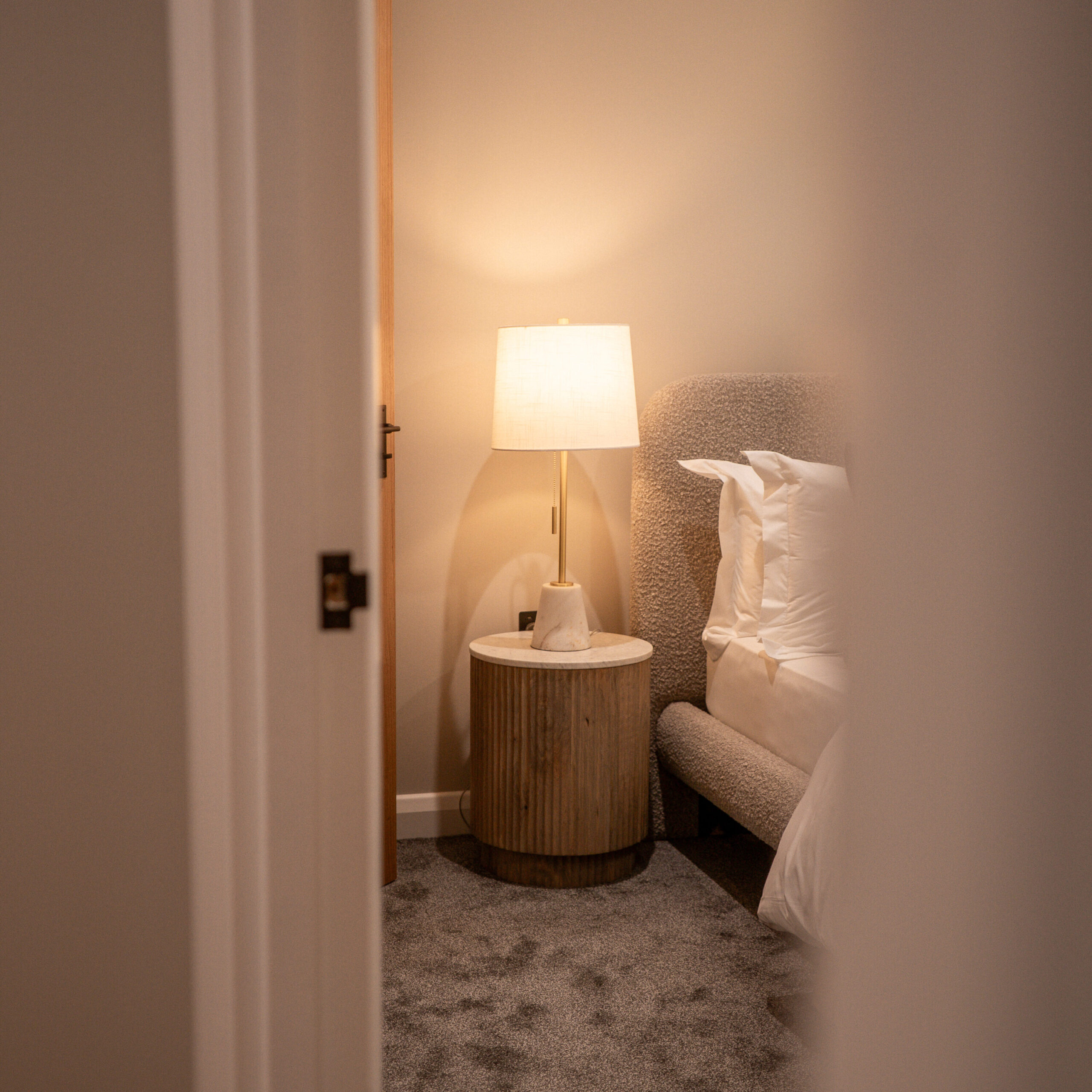 Looking from the hallway into the main bedroom, looking at warm light lamp on the bedside table. Next to boucle grey bed and white pillows