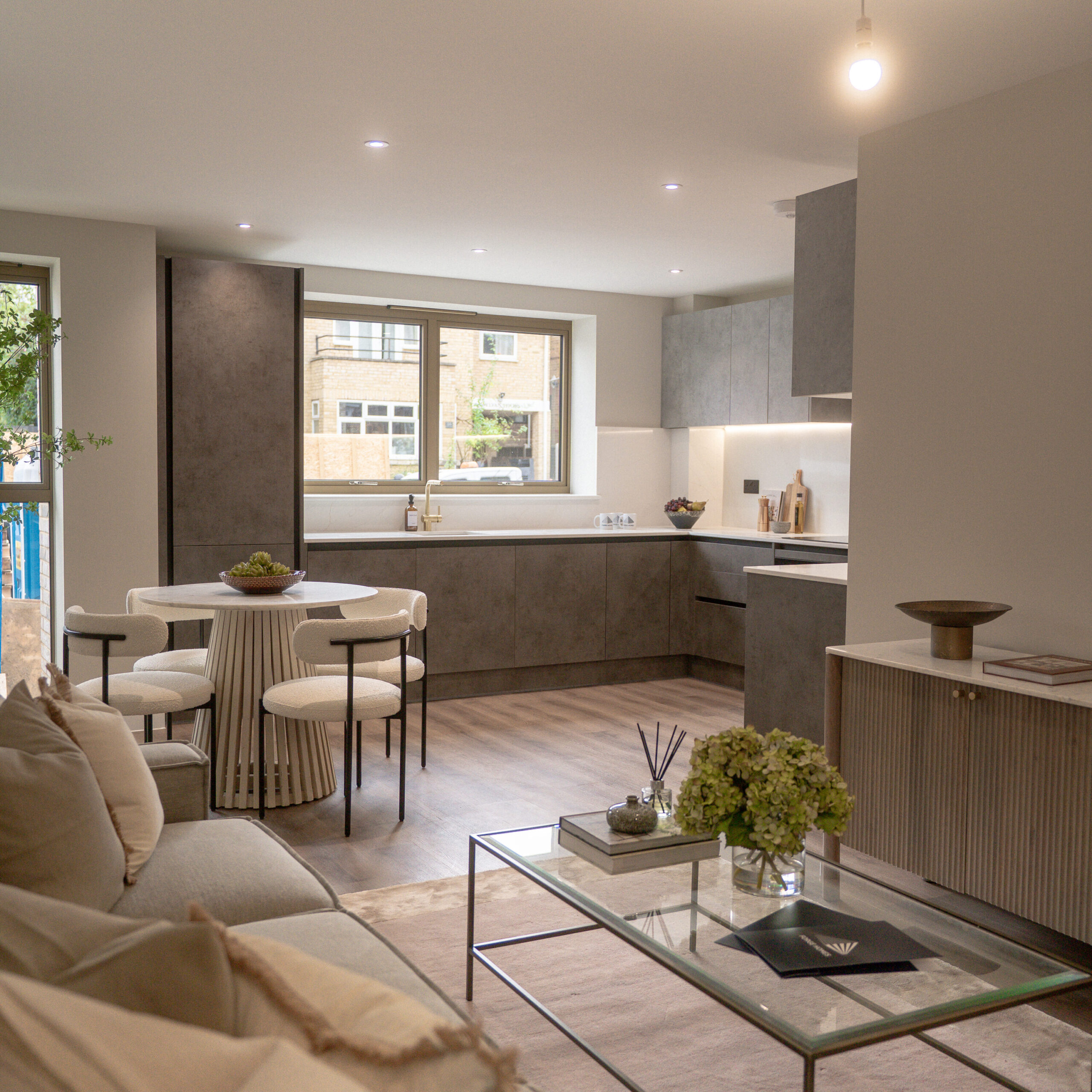 Living room, with gold exterior windows and grey sofas. Glass coffee table with more decorative pieces on top. Large purple tone rug underneath the sofa and table. Decorative artificial tree in the corner with dining table. Kitchen in the background with slate grey cabinets and marble countertops